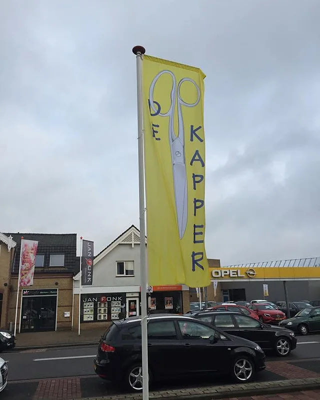 Gele vlag met een schaar en de woorden De Kapper, aan de straat